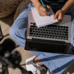Person taking notes while studying online using a laptop and notebook indoors